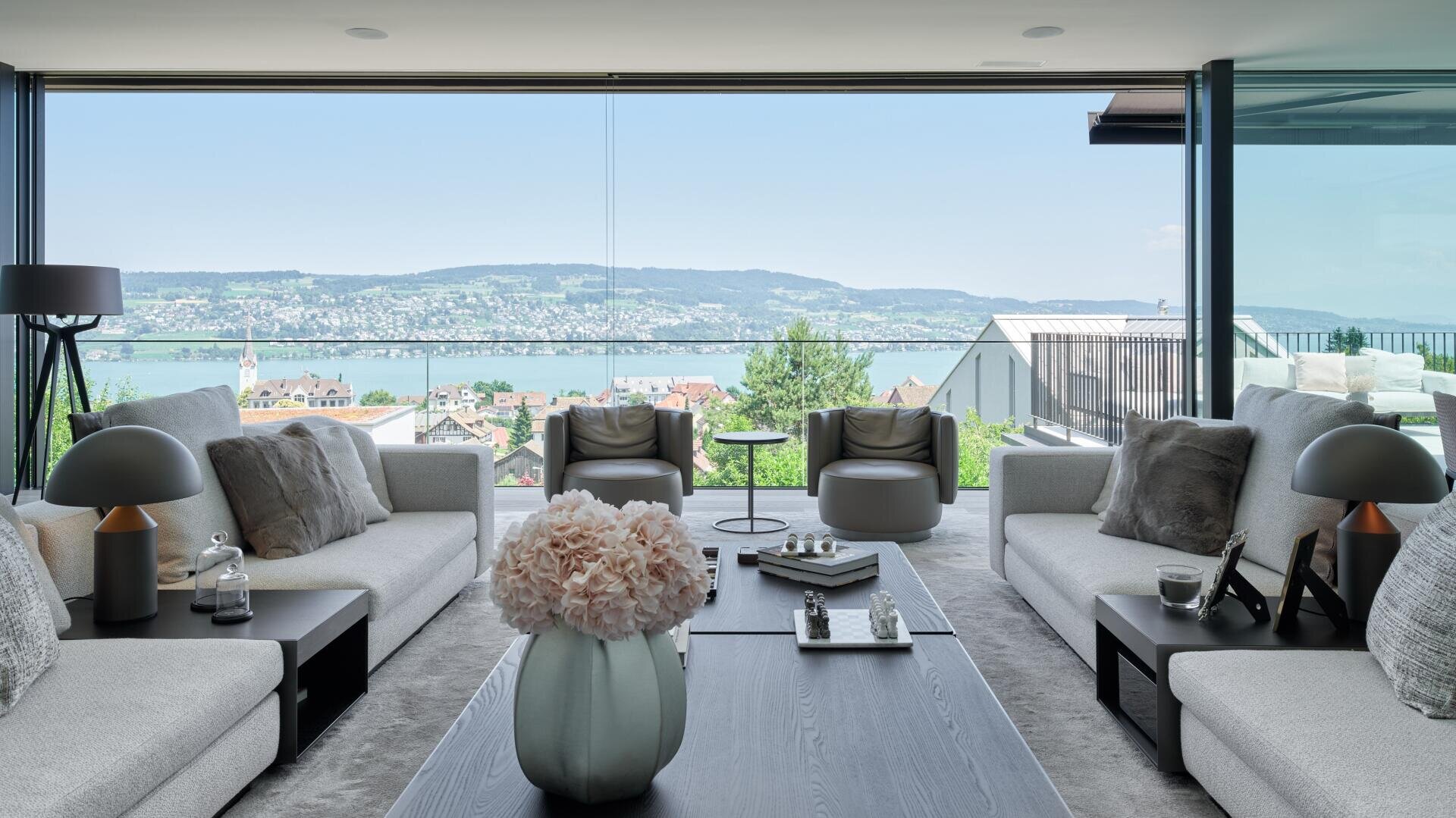 Ein modernes Wohnzimmer mit großen Glasfenstern bietet einen Panoramablick auf einen See, die Stadt und die Hügel. Der Raum verfügt über Sofas in neutralen Farben, Sessel, einen Couchtisch mit Büchern und dekorative Vasen mit Blumen.