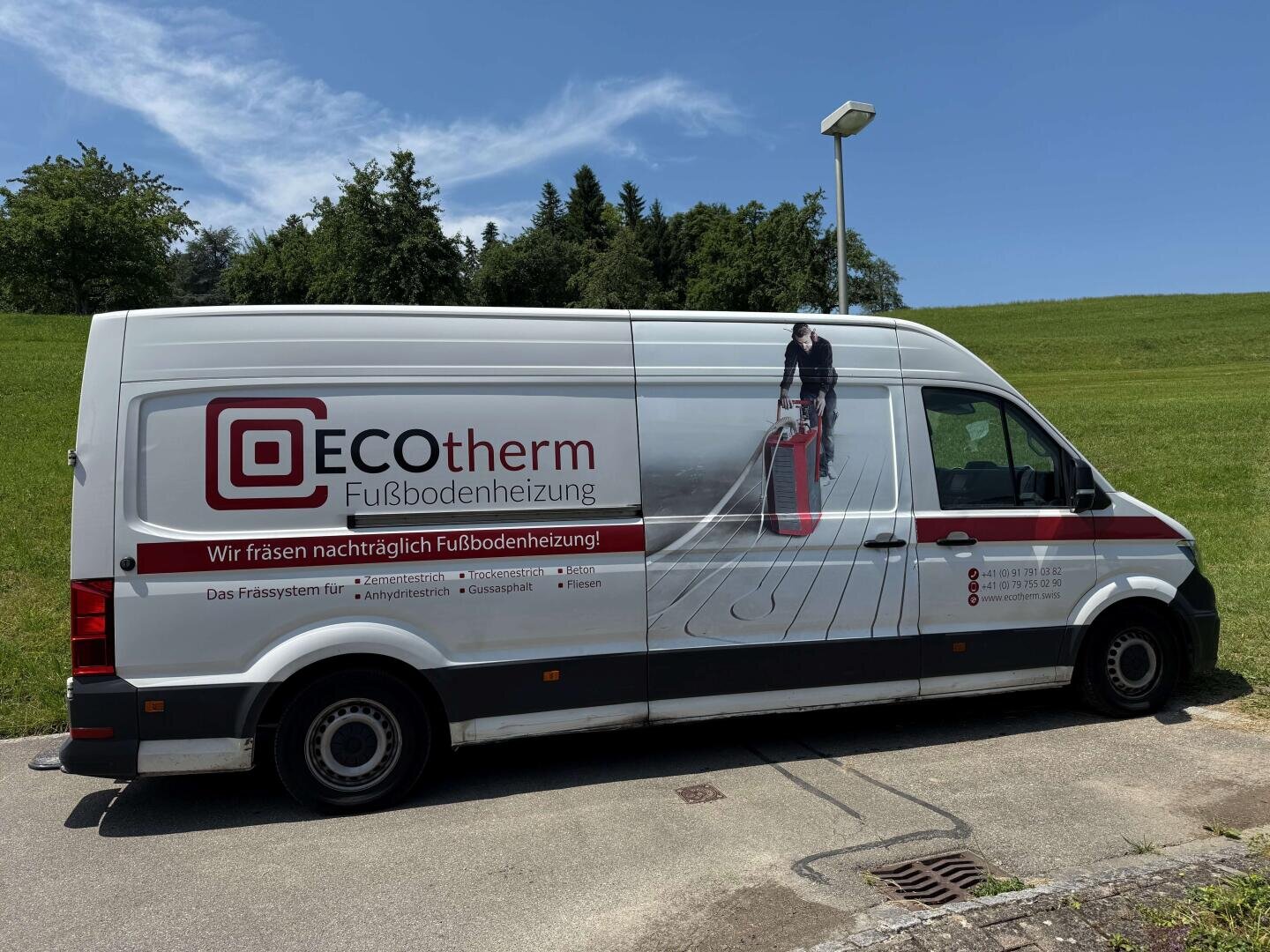 Ein weißer Lieferwagen mit der Aufschrift ECOtherm Fußbodenheizung ist am Straßenrand geparkt. Der Lieferwagen zeigt das Bild eines Arbeiters, der eine Fußbodenheizung benutzt, mit grünem Gras und Bäumen im Hintergrund.