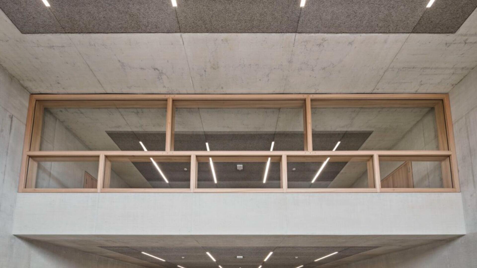 Ein modernes Interieur mit Betondecken und -wänden und rechteckigen, holzgerahmten Fenstern mit Blick auf einen Korridor. Lineare Deckenleuchten und Akustikplatten unterstreichen den minimalistischen architektonischen Stil.