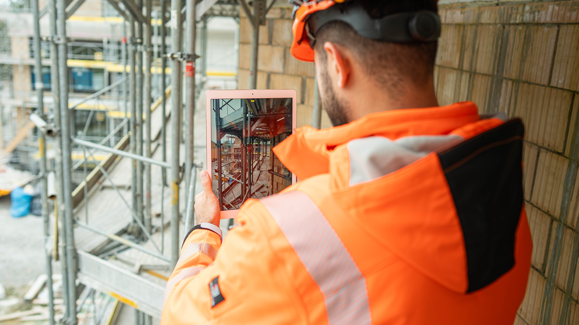 Ein Bauarbeiter in einer orangefarbenen Schutzjacke und mit Helm benutzt ein Tablet auf einer Baustelle mit Gerüsten und unfertigen Gebäuden im Hintergrund.