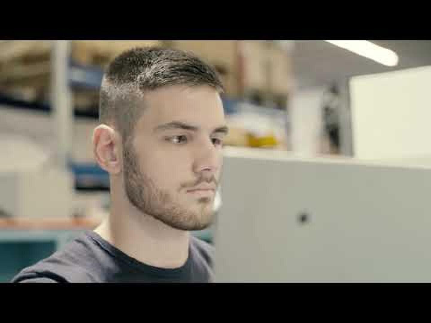 Ein junger Mann mit kurzen Haaren und Bart konzentriert sich auf einen Computerbildschirm in einem Innenraum, möglicherweise in einem Büro oder einer Werkstatt, wobei im unscharfen Hintergrund Regale und Geräte zu sehen sind.