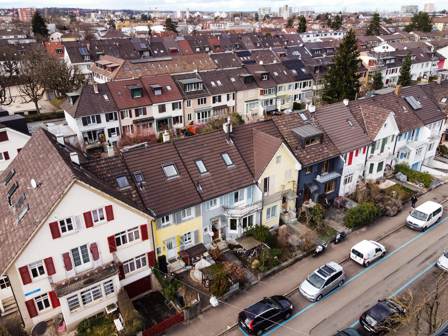 Luftaufnahme einer mit bunten Reihenhäusern, geparkten Autos und kleinen Vorgärten gesäumten Wohnstraße. Im Hintergrund sind bei bewölktem Himmel weitere Häuser und Gebäude zu sehen.