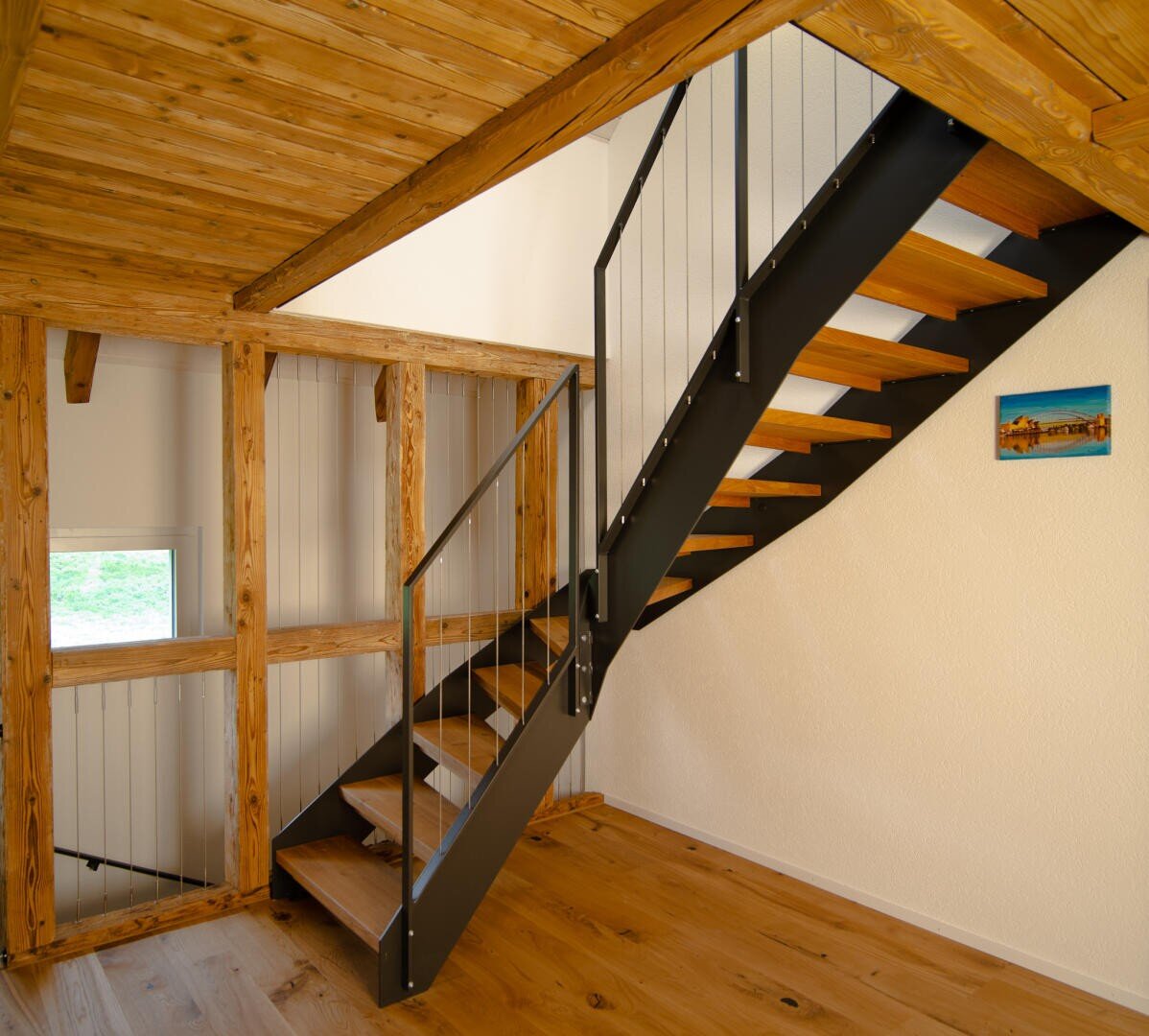 Escalier moderne en bois avec une rampe en métal noir dans un intérieur de style loft avec des poutres apparentes, des sols en bois clair, une petite fenêtre et un tableau coloré sur le mur couleur crème.