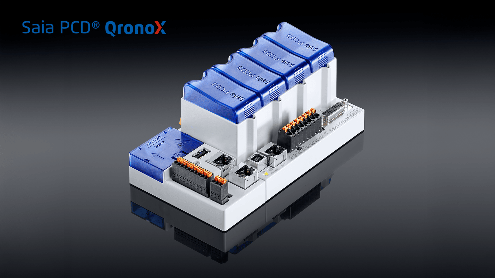 Eine programmierbare Saia PCD QronoX-Automatisierungssteuerung mit blauen und weissen Modulen, verschiedenen Ports und Anschlüssen, dargestellt auf einer reflektierenden schwarzen Oberfläche.