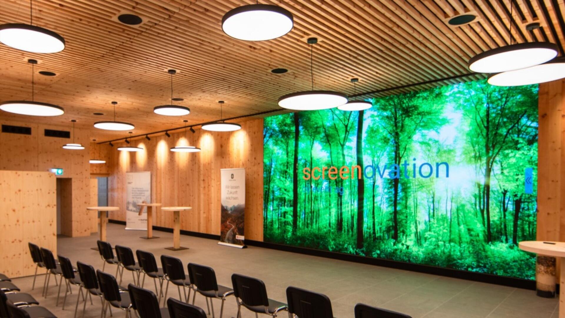 Ein moderner Konferenzraum mit hölzernen Wänden und Decken, schwarzen Stuhlreihen, hohen runden Tischen und einem großen digitalen Bildschirm, auf dem eine helle Waldszene mit dem Wort Screenovation zu sehen ist.