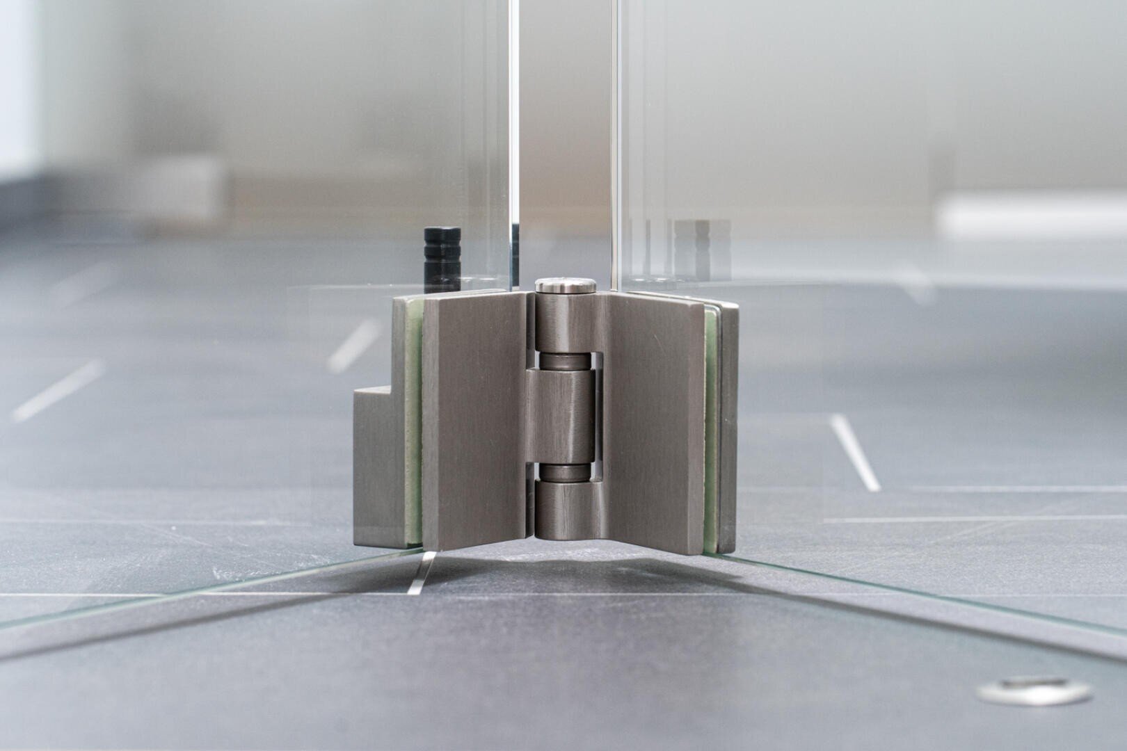Nahaufnahme eines Metallscharniers, das zwei Glasscheiben auf einem grau gefliesten Boden verbindet, wahrscheinlich Teil einer Duschtür oder einer Glastrennwand in einer modernen Inneneinrichtung.