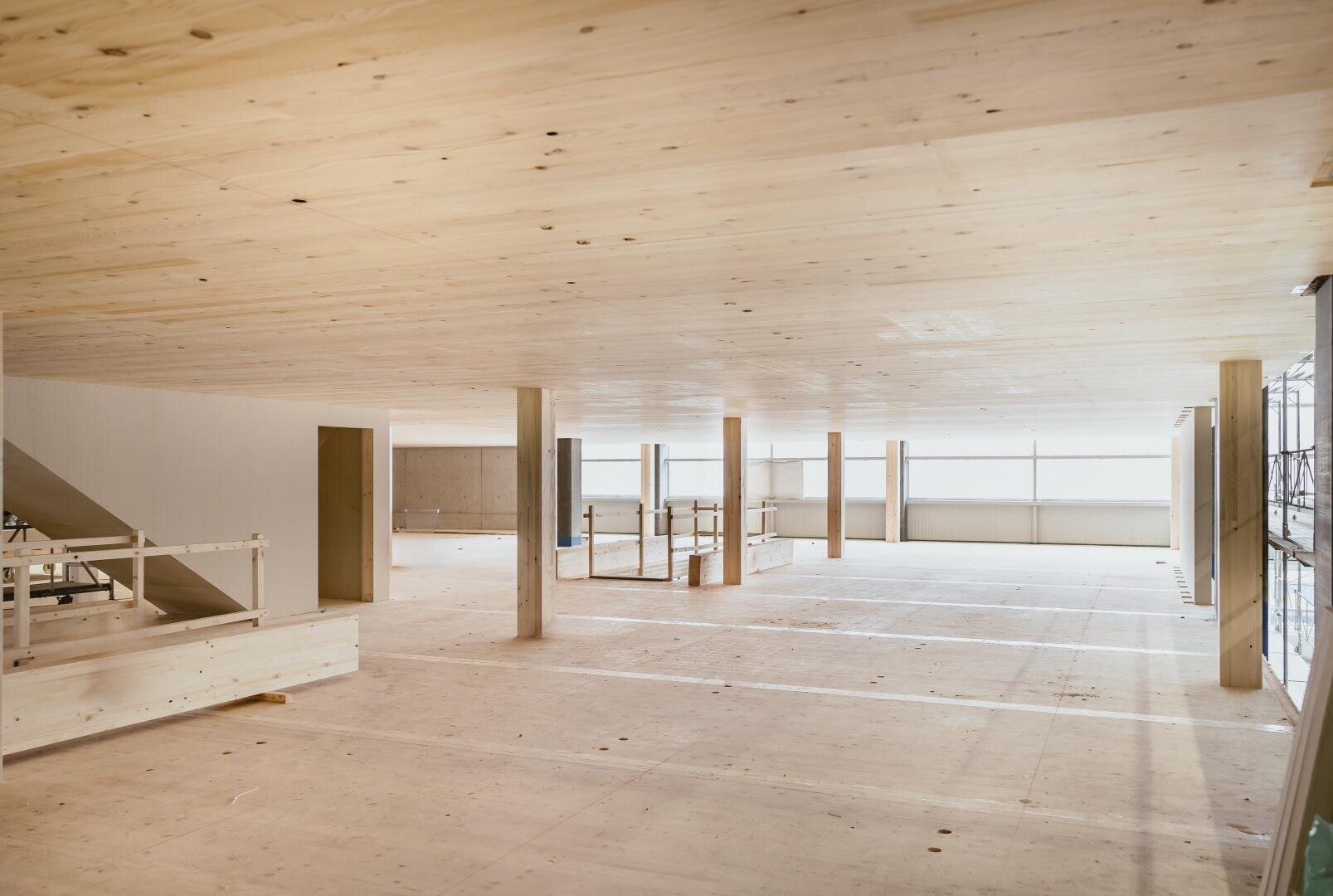 Ein geräumiger, unfertiger Innenraum mit hellen Holzböden, Decken und Stützpfeilern. Große Fenster säumen eine Wand und lassen reichlich natürliches Licht herein. Auf der linken Seite sind Baumaterialien und ein Treppenhaus zu sehen.