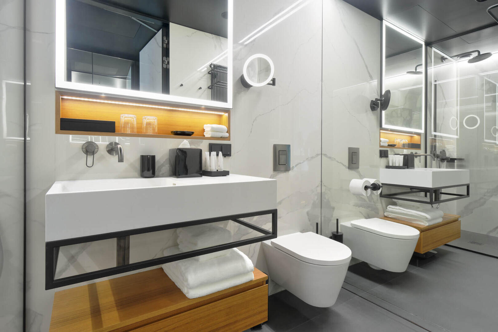 Modernes Bad mit schwimmendem Waschbecken und Toilette, Holzregalen für Handtücher, großen Spiegeln, wandmontierten Armaturen und einem beleuchteten Spiegel über dem Waschbecken. Der Raum ist in schlichtem, minimalistischem Weiß und Grau gehalten.