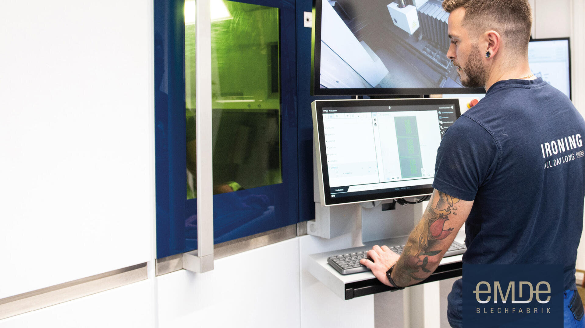 Ein Mann bedient einen Computer an einem Industriearbeitsplatz, mit Monitoren und Maschinen im Hintergrund. Er trägt ein blaues Hemd mit der Aufschrift "IRIONING" auf dem Rücken, und in der rechten unteren Ecke ist das Logo "eMDe BLECHFABRIK" zu sehen.