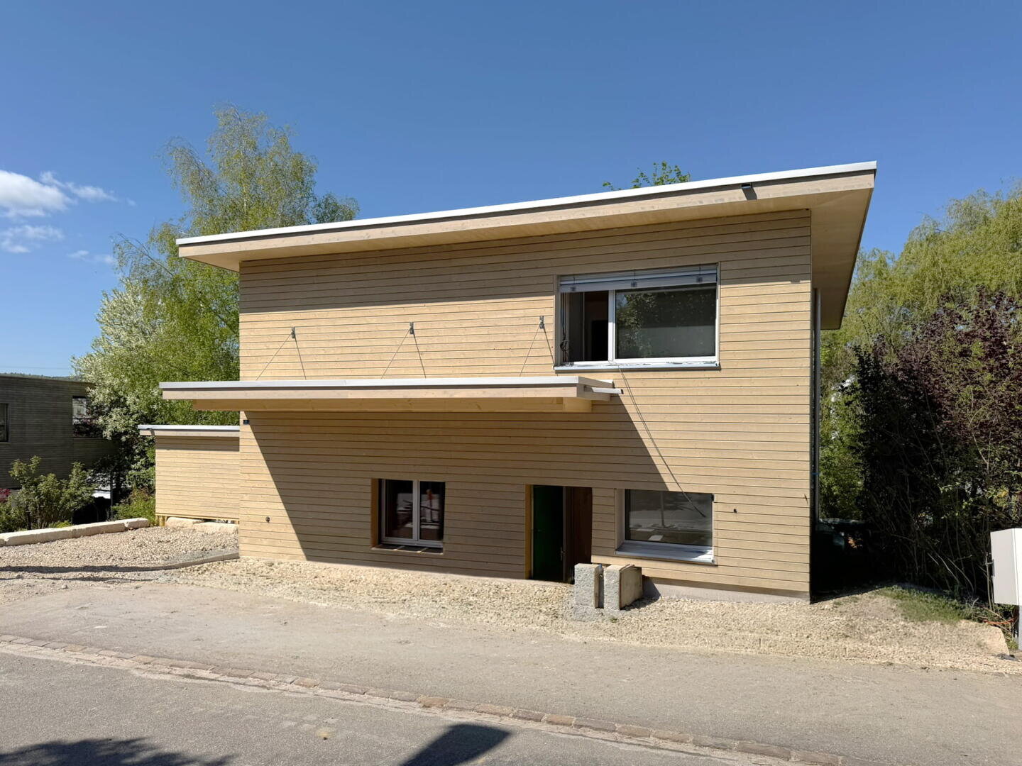 Ein modernes zweistöckiges Haus mit hellen Holzverkleidungen, einem Flachdach und großen Fenstern. Der Hof ist unbearbeitet und mit Kies bedeckt, im Hintergrund stehen Bäume und Sträucher unter einem klaren blauen Himmel.