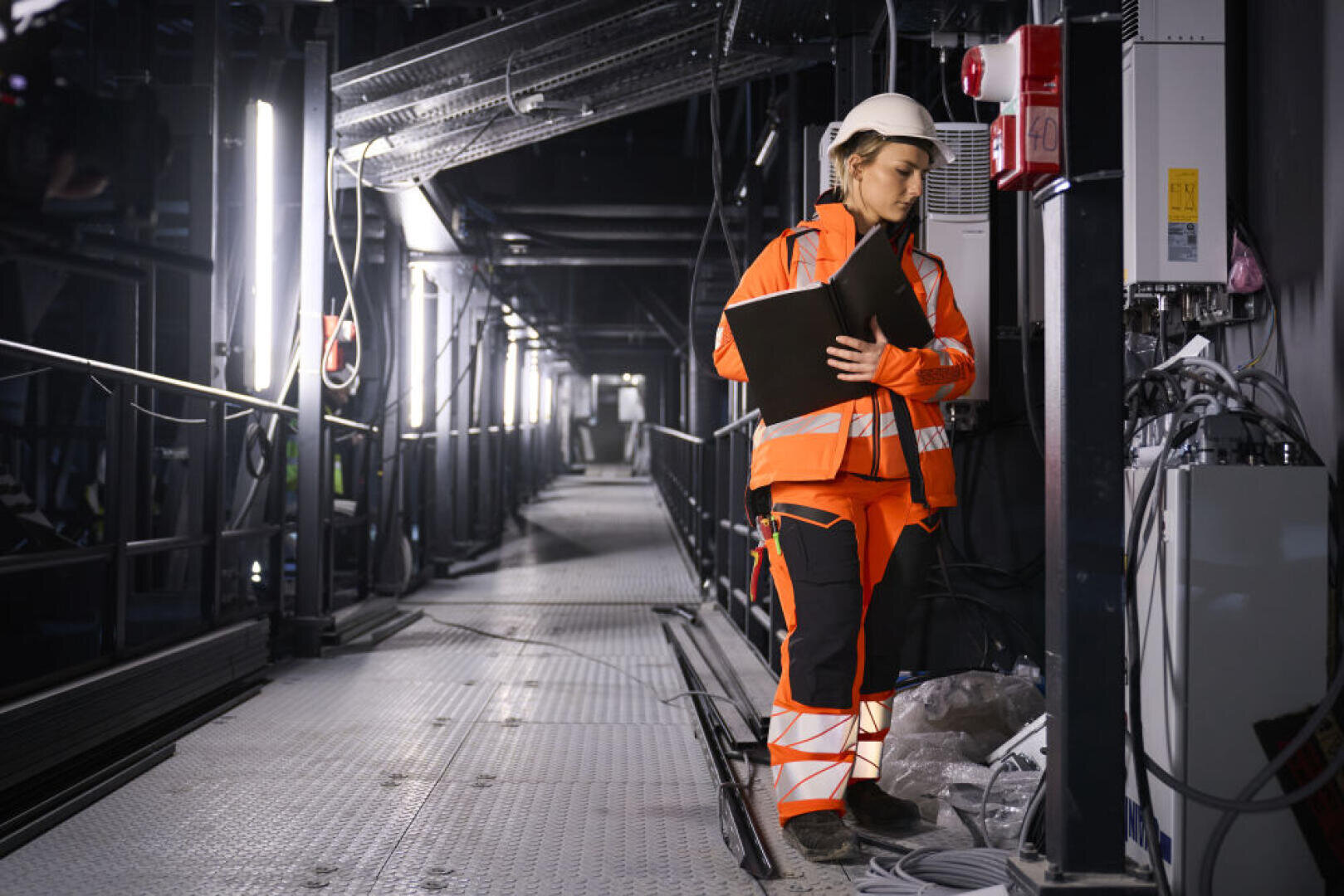Une personne portant une veste de sécurité orange, un pantalon et un casque de protection blanc tient une planchette à pince pendant qu'elle vérifie l'équipement dans un environnement industriel ou de construction avec des passerelles métalliques et un éclairage au plafond.