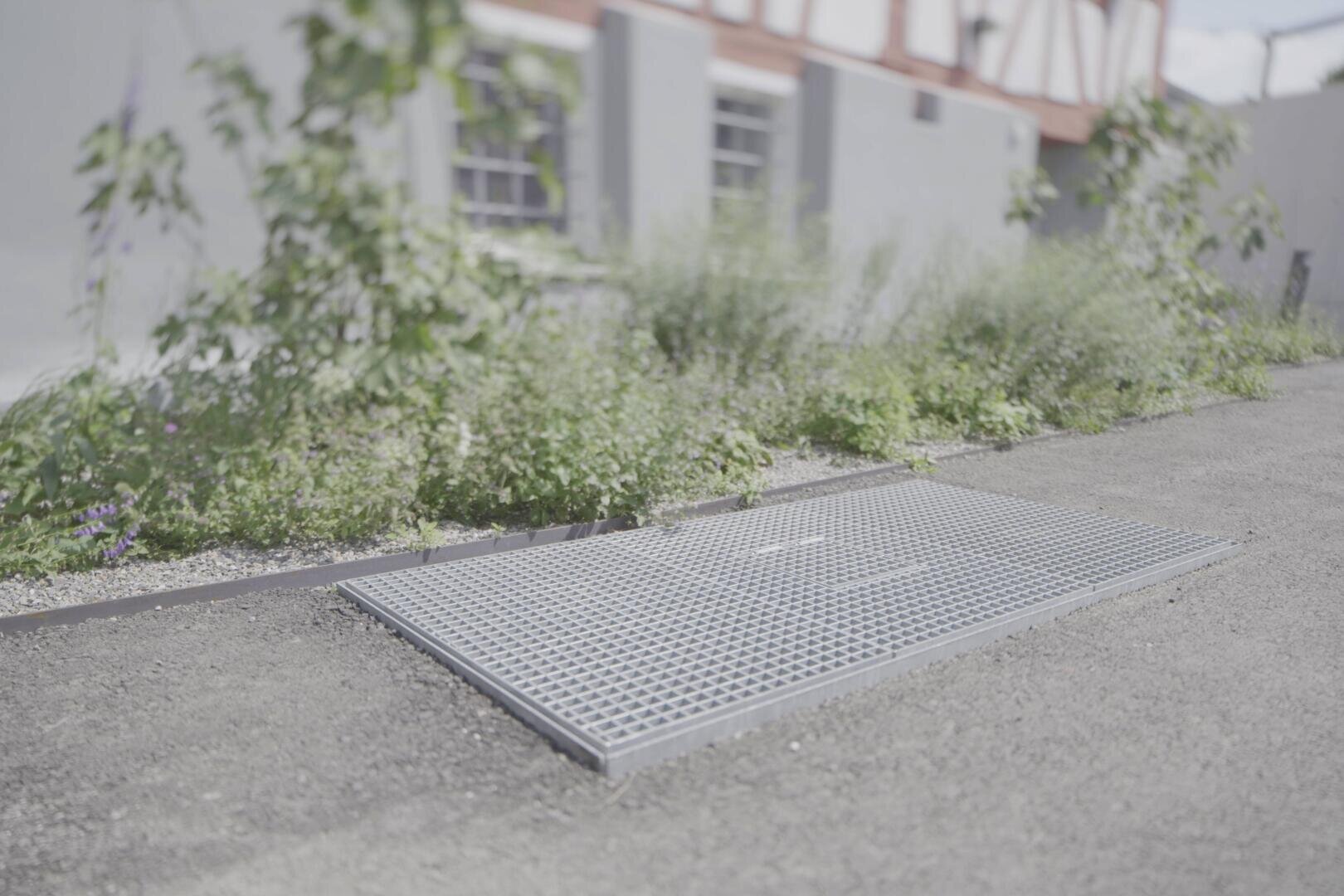 Une grille métallique encastrée dans une allée pavée à côté d'un petit jardin avec des plantes vertes, devant un bâtiment clair avec des fenêtres.