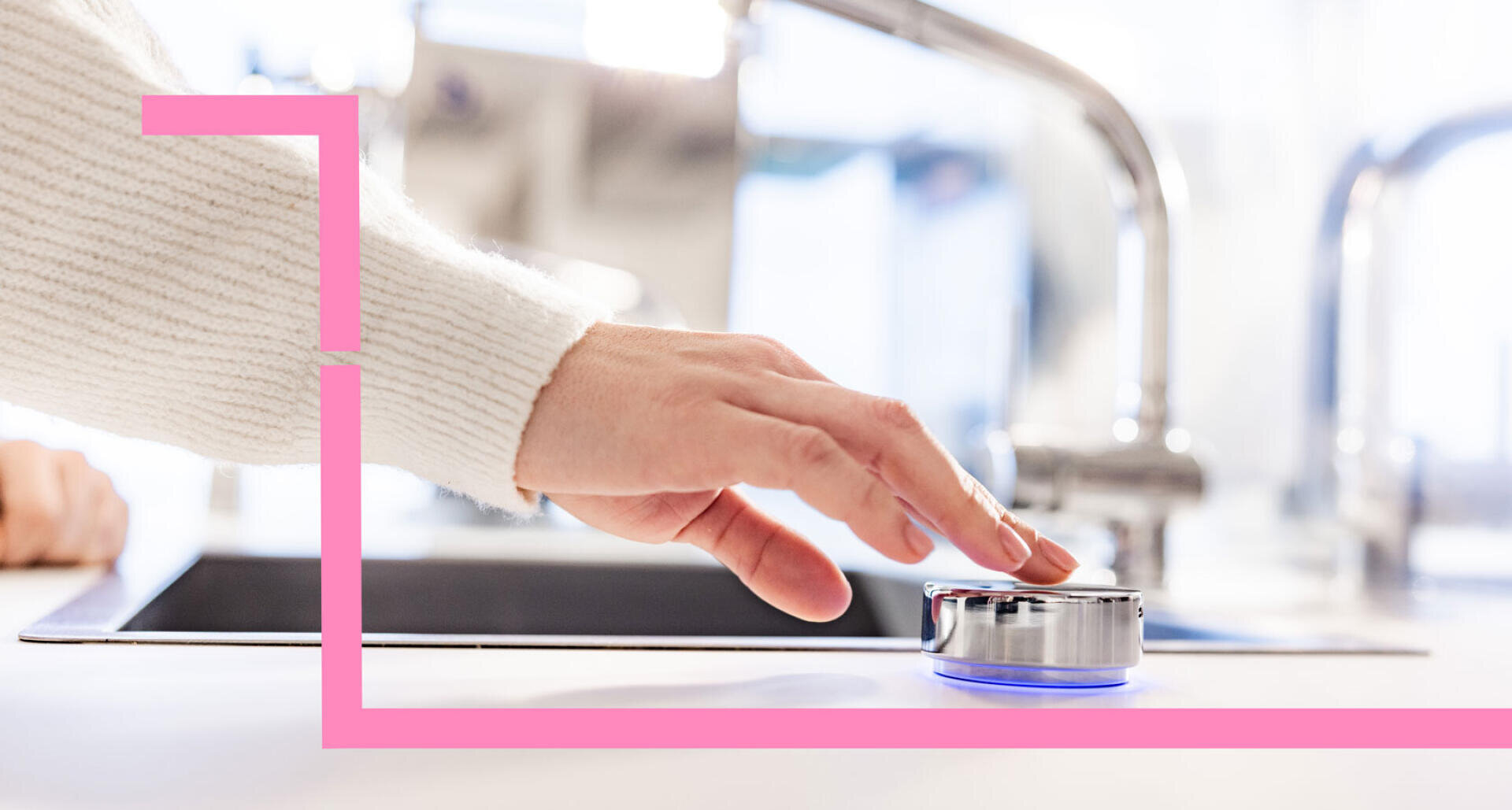 Une personne portant un pull blanc touche un appareil intelligent rond et métallique sur un comptoir de cuisine blanc, avec un évier et un robinet flous en arrière-plan. Une ligne graphique rose encadre une partie de l'image.