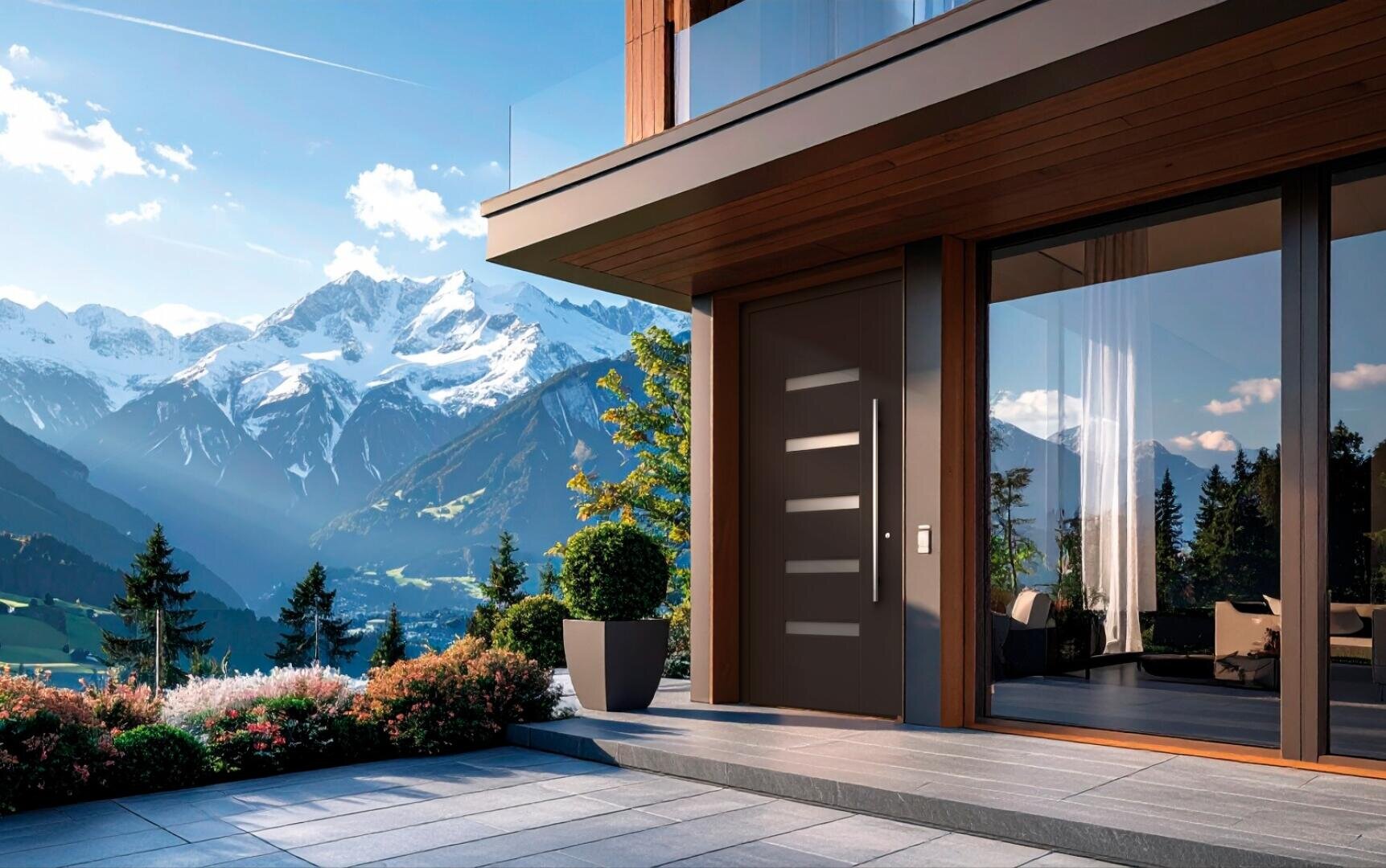 Une maison moderne avec de grandes fenêtres et une porte d'entrée élégante se dresse sur une terrasse carrelée donnant sur une verdure luxuriante et de grands pins, avec des montagnes enneigées en arrière-plan sous un ciel bleu limpide.
