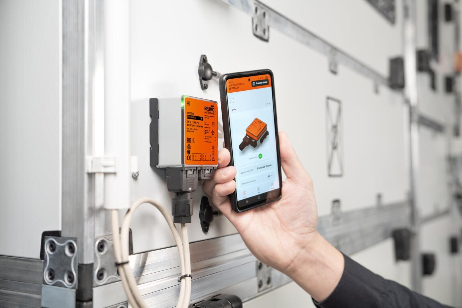 Eine Person hält ein Smartphone in der Hand, das Informationen über ein orangefarbenes elektronisches Gerät anzeigt, das auf einer weißen Industrietafel montiert und mit Kabeln an das Gerät angeschlossen ist.