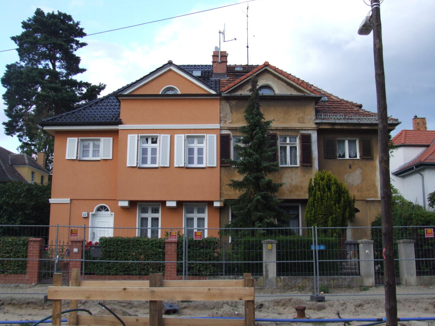 Ein in zwei Hälften geteiltes Haus, wobei die linke Seite renoviert und orangefarben mit weißer Zierleiste gestrichen ist, während die rechte Seite alt, verwittert und ungestrichen erscheint. Davor sind ein Zaun und Baumaterialien zu sehen.