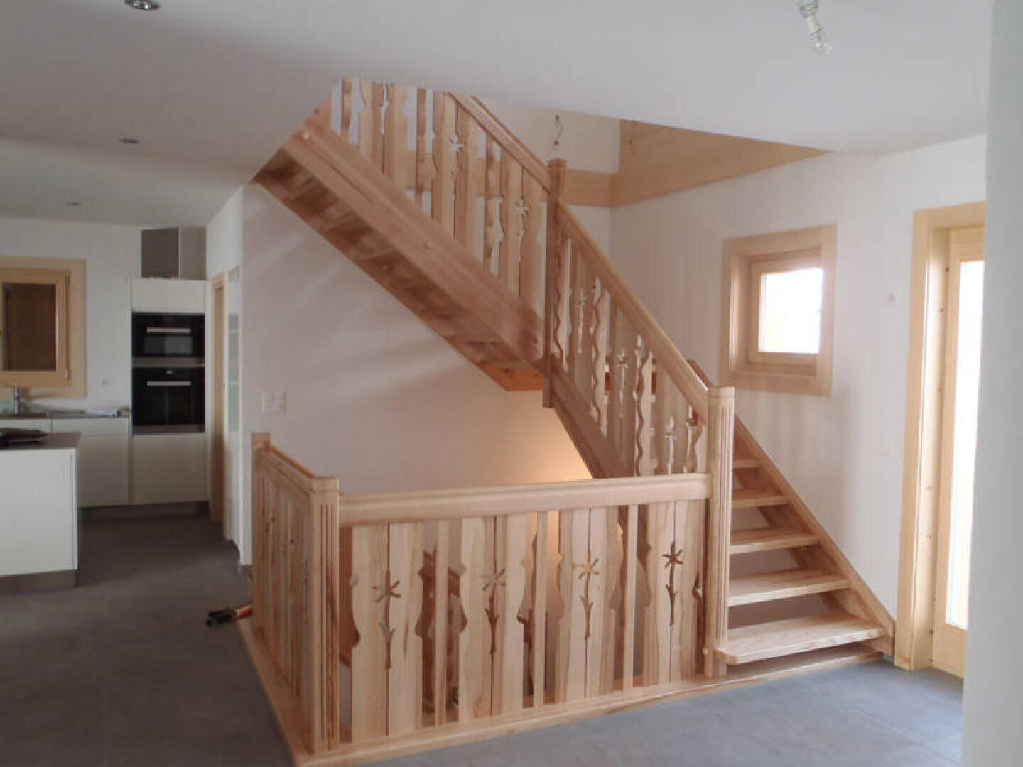 Un escalier en bois avec des rampes décoratives et des balustres dans un intérieur moderne et lumineux. L'escalier mène à un étage supérieur et est entouré de murs blancs, de boiseries claires et d'un sol carrelé.