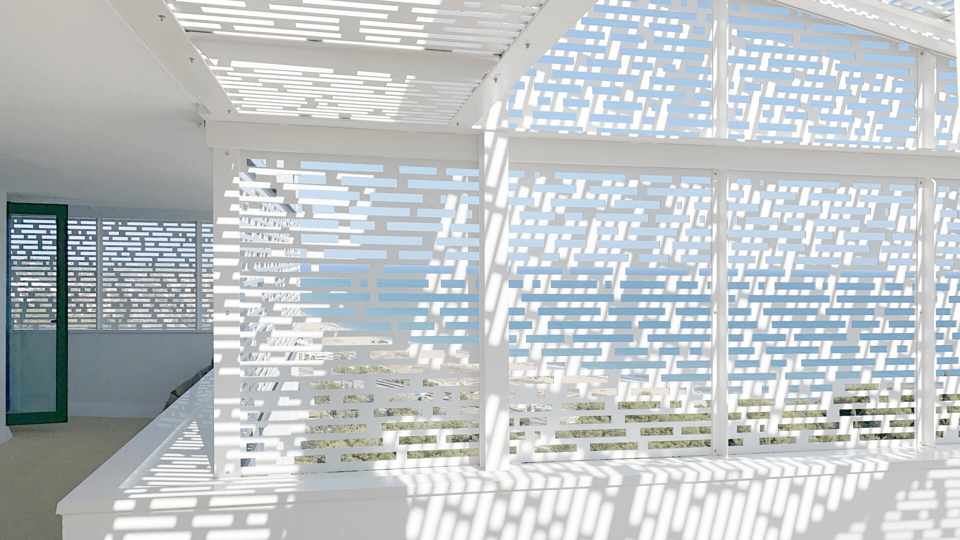 Ein heller Balkon mit weißen, geometrischen Schirmen, die gemusterte Schatten werfen, mit Blick auf das blaue Meer und die Küstenlinie unter einem klaren Himmel.