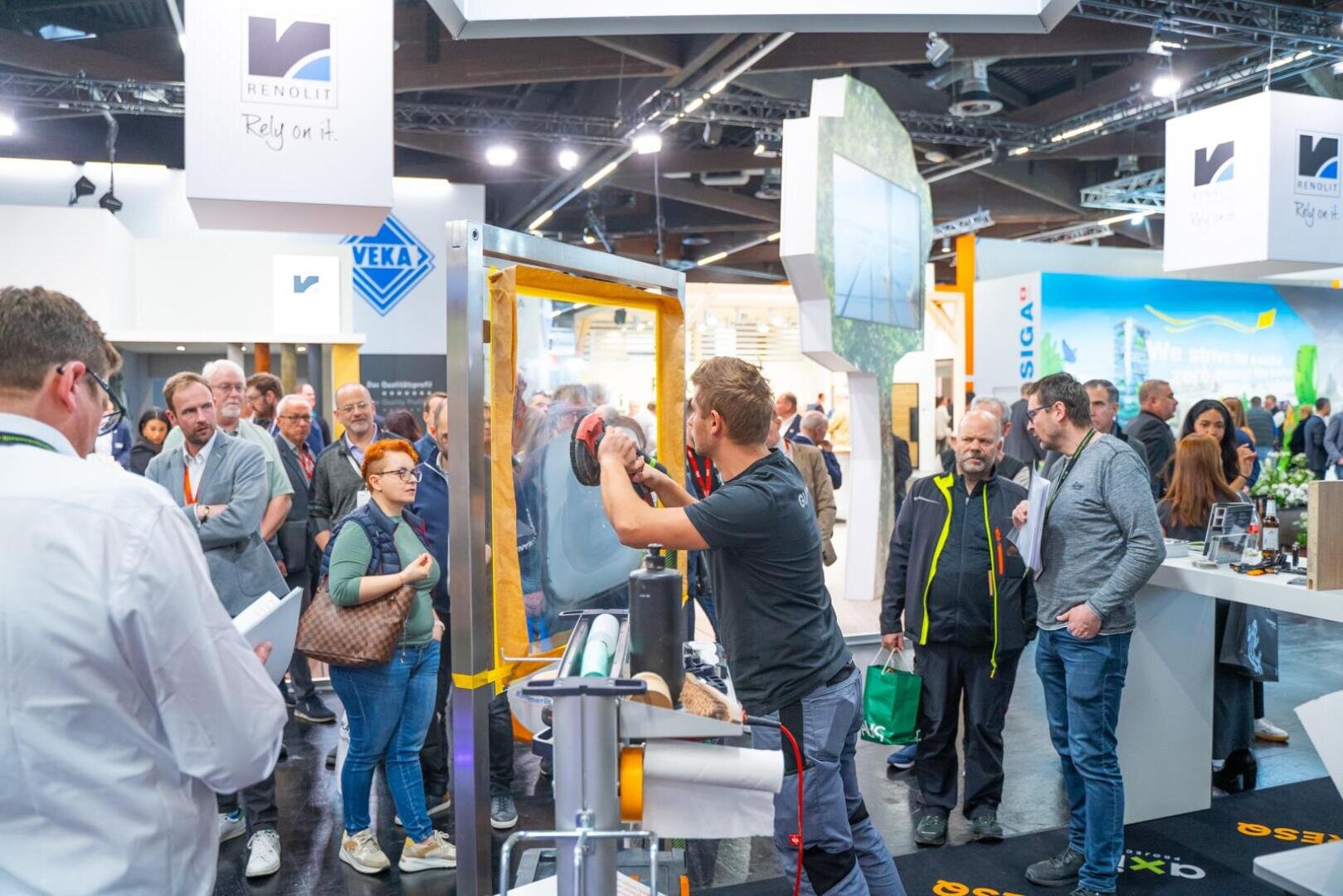 Ein Mann demonstriert einer Gruppe von Menschen auf einer belebten Messe in einer Halle ein Werkzeug an einem Fensterrahmen. Die Teilnehmer schauen aufmerksam zu, während im Hintergrund Firmenbanner und Messestände zu sehen sind.