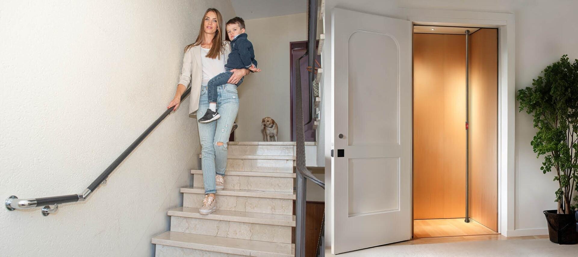 Une femme porte un petit enfant en bas d'un escalier avec une main courante, tandis qu'un petit chien attend en haut ; à côté, on voit un ascenseur d'immeuble ouvert avec des murs en boiseries.