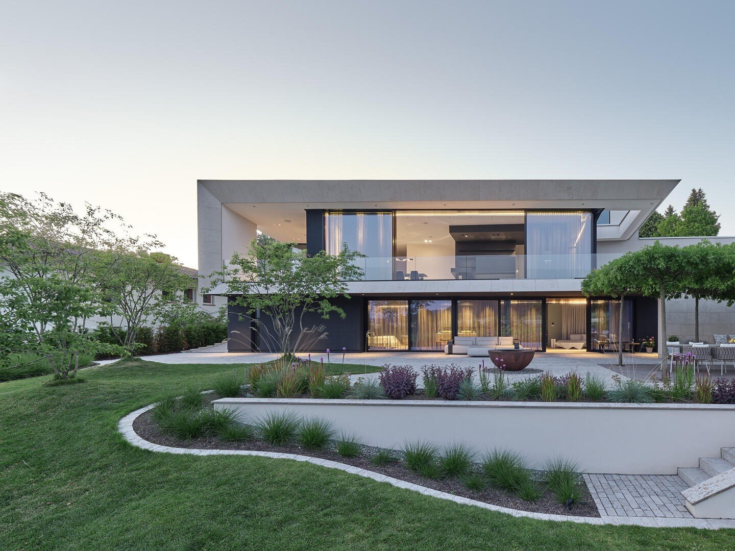 Une maison moderne de deux étages avec de grandes baies vitrées, entourée d'un jardin aménagé avec de l'herbe verte, des arbres et des plantes bien disposées, photographiée au crépuscule, les lumières étant visibles à l'intérieur de la maison.