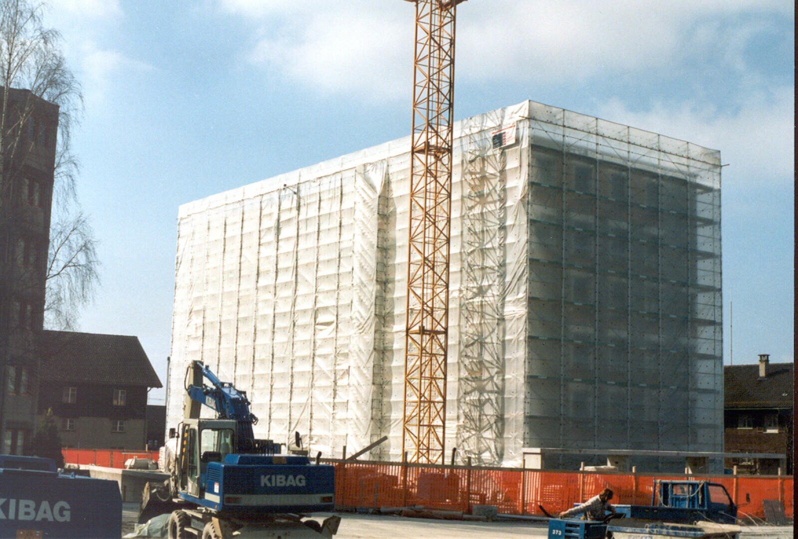 Ein großes, im Bau befindliches Gebäude ist mit weißen Gerüsten und Schutznetzen bedeckt. Davor steht ein großer gelber Kran, auf dem Boden liegen Baumaschinen und Materialien. Der Himmel ist teilweise bewölkt.