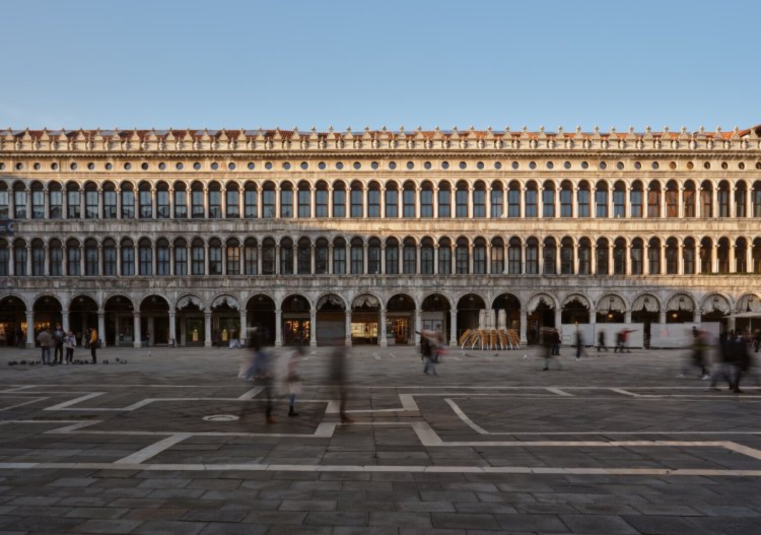Auf dem Markusplatz in Venedig (Italien) ist ein großes historisches Gebäude mit Reihen von Bögen und Säulen bei Sonnenuntergang zu sehen. Menschen gehen unscharf über den offenen Steinplatz davor.
