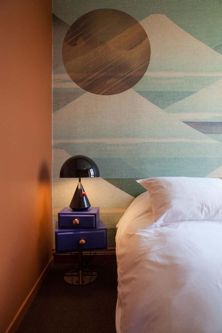 Une chambre à coucher moderne avec un lit blanc, une table de nuit bleue à deux tiroirs, une lampe noire et un mur décoratif avec des montagnes abstraites et un grand cercle. Un éclairage chaud et un mur orange sont également visibles.