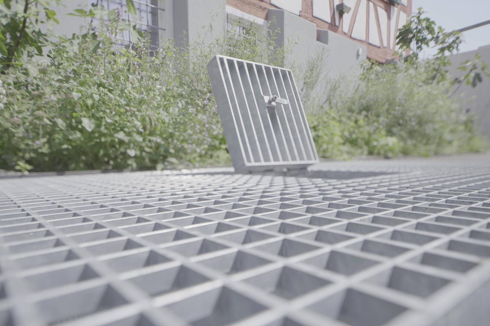 Gros plan sur une plateforme en treillis métallique à l'extérieur, dont une grille carrée est ouverte en guise de trappe ; en arrière-plan, on voit des plantes vertes et un bâtiment blanc.