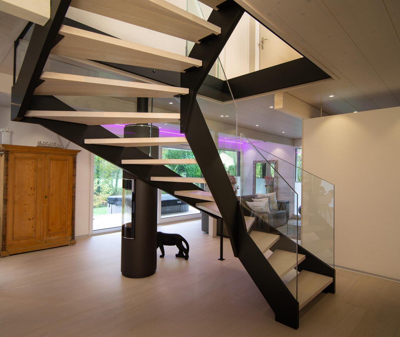 Un intérieur moderne avec un escalier flottant avec des supports métalliques noirs et des marches en bois clair, des balustrades en verre, une grande fenêtre, un éclairage d'accentuation violet, une armoire en bois et une sculpture de chien noire sous l'escalier.