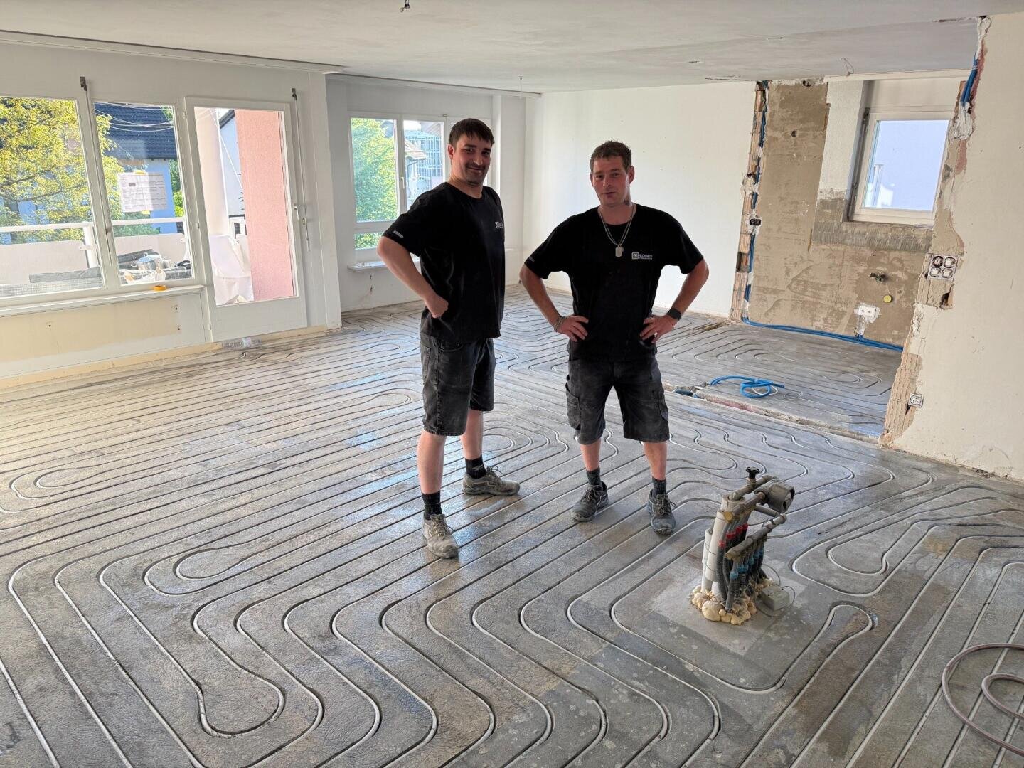 Zwei Männer in Arbeitskleidung stehen in einem hellen, teilweise renovierten Raum mit im Betonboden verlegten Fußbodenheizungsrohren. Im Hintergrund sind Baumaterialien und freigelegte Wände zu sehen.