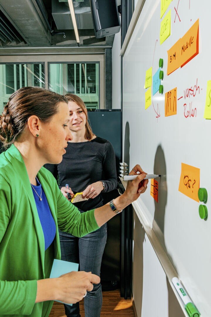 Zwei Frauen arbeiten gemeinsam an einem Whiteboard, das mit bunten Haftnotizen und Magneten bedeckt ist. Eine Frau schreibt mit einem Marker auf das Board, während die andere lächelnd zusieht und weitere Haftnotizen in der Hand hält.