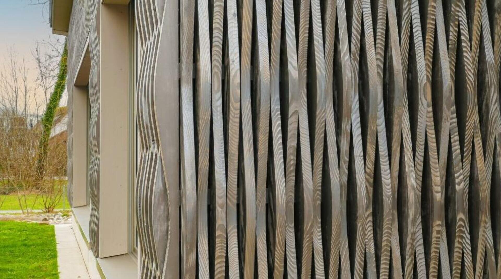 Nahaufnahme einer modernen Gebäudefassade mit vertikalen Holzlatten mit wellenförmigen Mustern, die eine strukturierte und dynamische Oberfläche schaffen; im Hintergrund sind grünes Gras und Bäume zu sehen.
