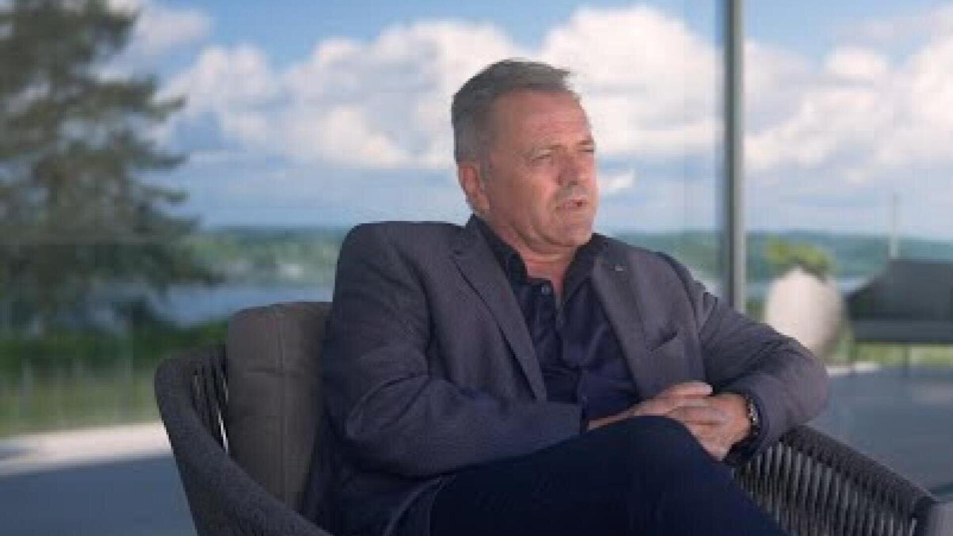 Un homme vêtu d'un blazer sombre est assis sur une chaise en plein air, les mains croisées, l'air pensif. Derrière lui, on voit un arrière-plan flou avec de la verdure, de l'eau et un ciel partiellement nuageux.