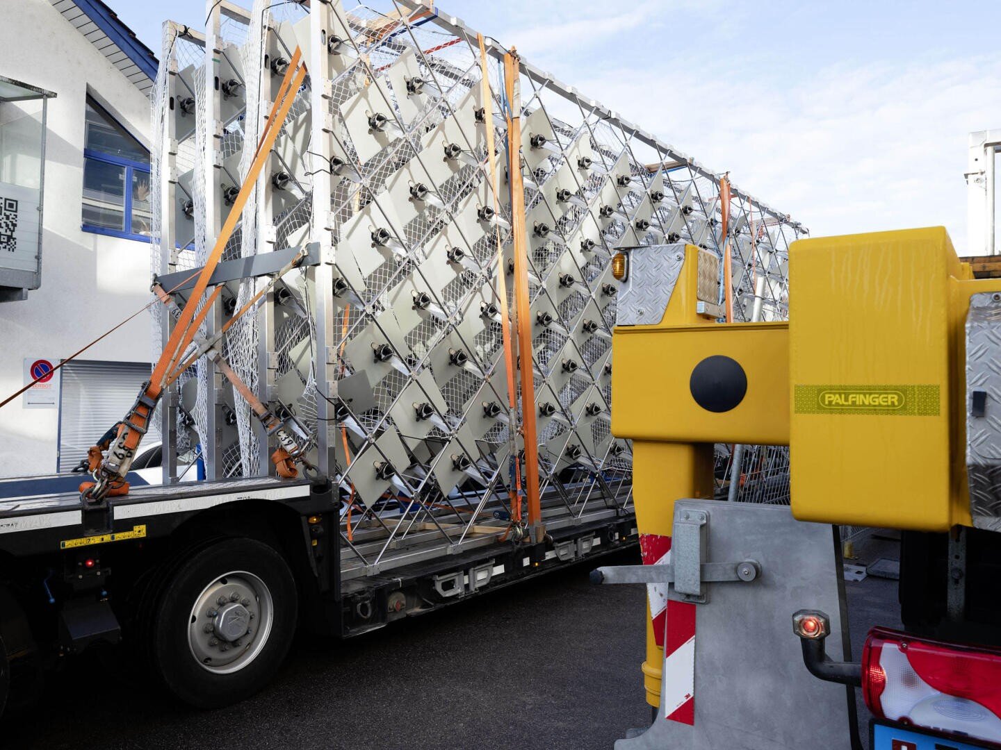 Une grande structure métallique avec plusieurs ventilateurs est fixée par des sangles orange sur un camion à plateau, à côté duquel se trouvent des machines jaunes portant l'inscription PALFINGER. La scène semble se dérouler dans une installation industrielle ou commerciale.