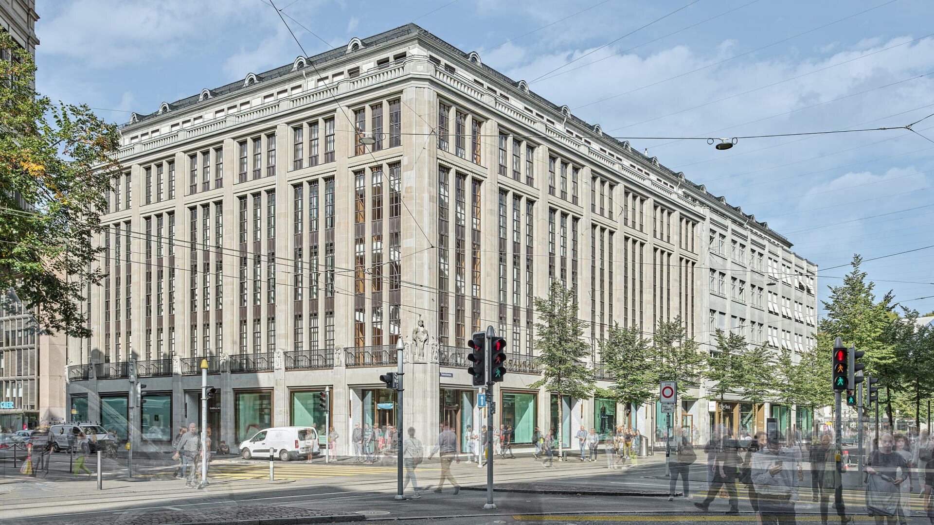 Ein großes, historisches, mehrstöckiges Gebäude mit vielen Fenstern steht an einer belebten Straßenkreuzung, an der Menschen die Straße überqueren, Autos geparkt sind und Bäume den Bürgersteig unter freiem Himmel säumen.