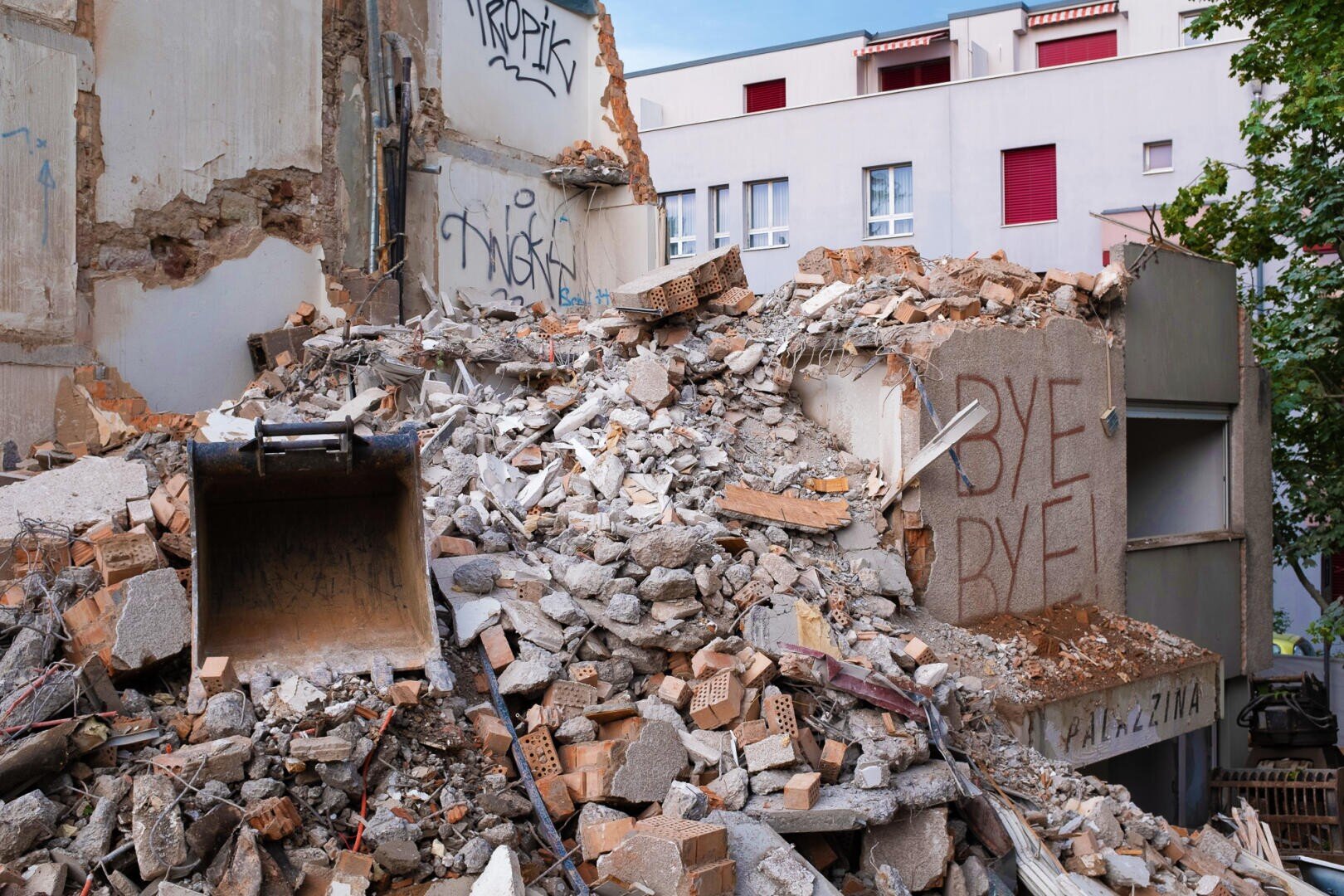 Ein Schutthaufen eines abgerissenen Gebäudes liegt vor teilweise noch stehenden Mauern mit Graffiti, darunter die Worte BYE BYE, die auf ein Stück Schutt geschrieben sind; im Hintergrund sind Wohnhäuser zu sehen.