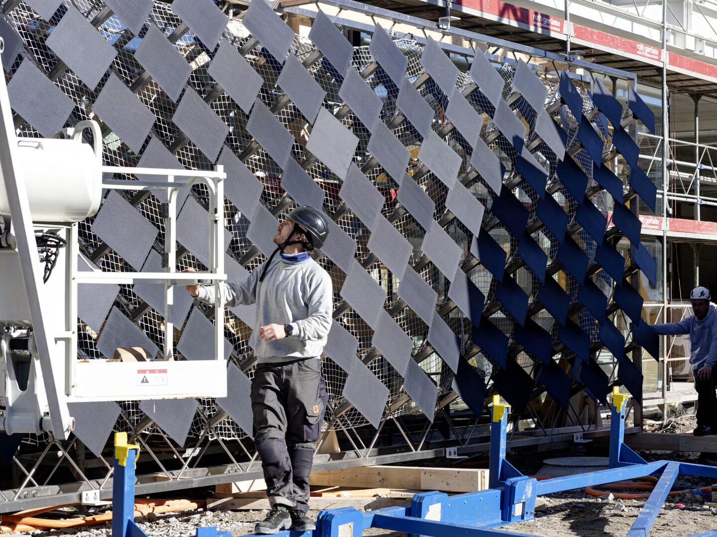 Un ouvrier du bâtiment portant un casque et un pull gris se tient à proximité d'un ascenseur et regarde une grande installation murale géométrique composée de panneaux en forme de losange. Un autre ouvrier se tient à l'arrière-plan. Derrière eux, on aperçoit un échafaudage.