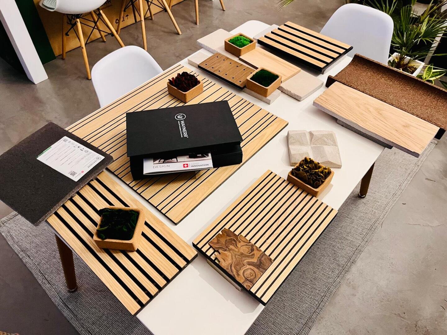 Une table avec des motifs en bois, une boîte noire, des petits plateaux avec de la mousse, des carreaux et des documents, entourés de chaises blanches dans une pièce moderne et bien éclairée.