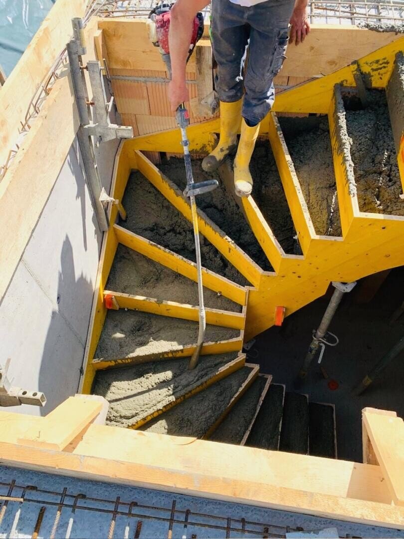 Ein Bauarbeiter in gelben Stiefeln glättet den nassen Beton einer geschwungenen Treppe mit einem Finishing Tool, umgeben von Holzschalungen und Stahlarmierungen.
