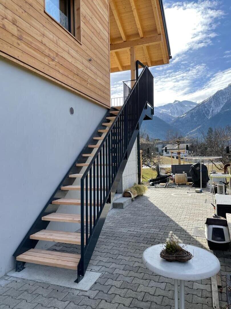 Un escalier extérieur moderne avec des marches en bois et des rampes en métal noir mène au balcon d'une maison en bois. Au premier plan, une table ronde blanche est posée sur un patio pavé avec des montagnes visibles à l'arrière-plan.