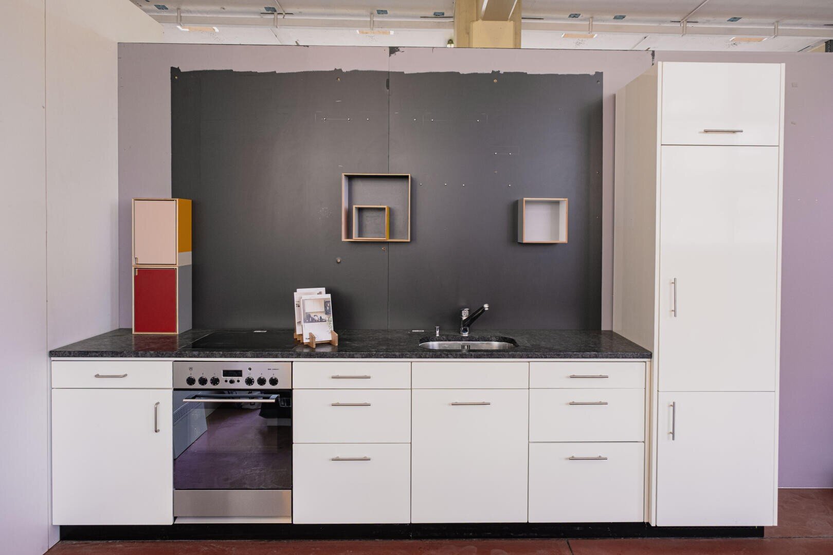 Cuisine moderne avec des armoires blanches, un plan de travail noir, un four encastré, un évier en acier inoxydable et des étagères géométriques sur un mur d'accent sombre. La cuisine donne une impression de minimalisme et d'ordre.