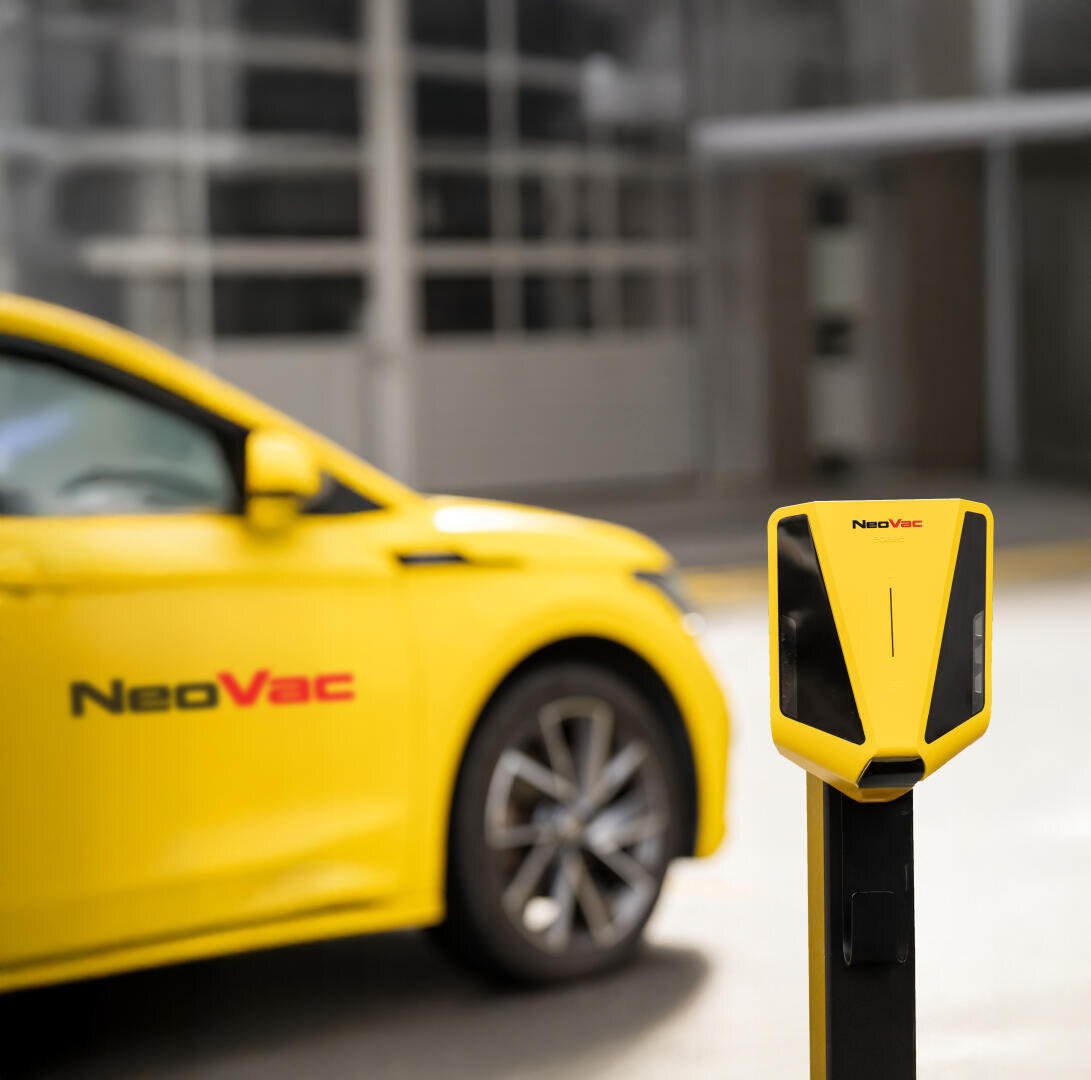 Une voiture électrique jaune de la marque NeoVac est garée à côté d'une station de recharge NeoVac jaune assortie, avec un bâtiment flou en arrière-plan.
