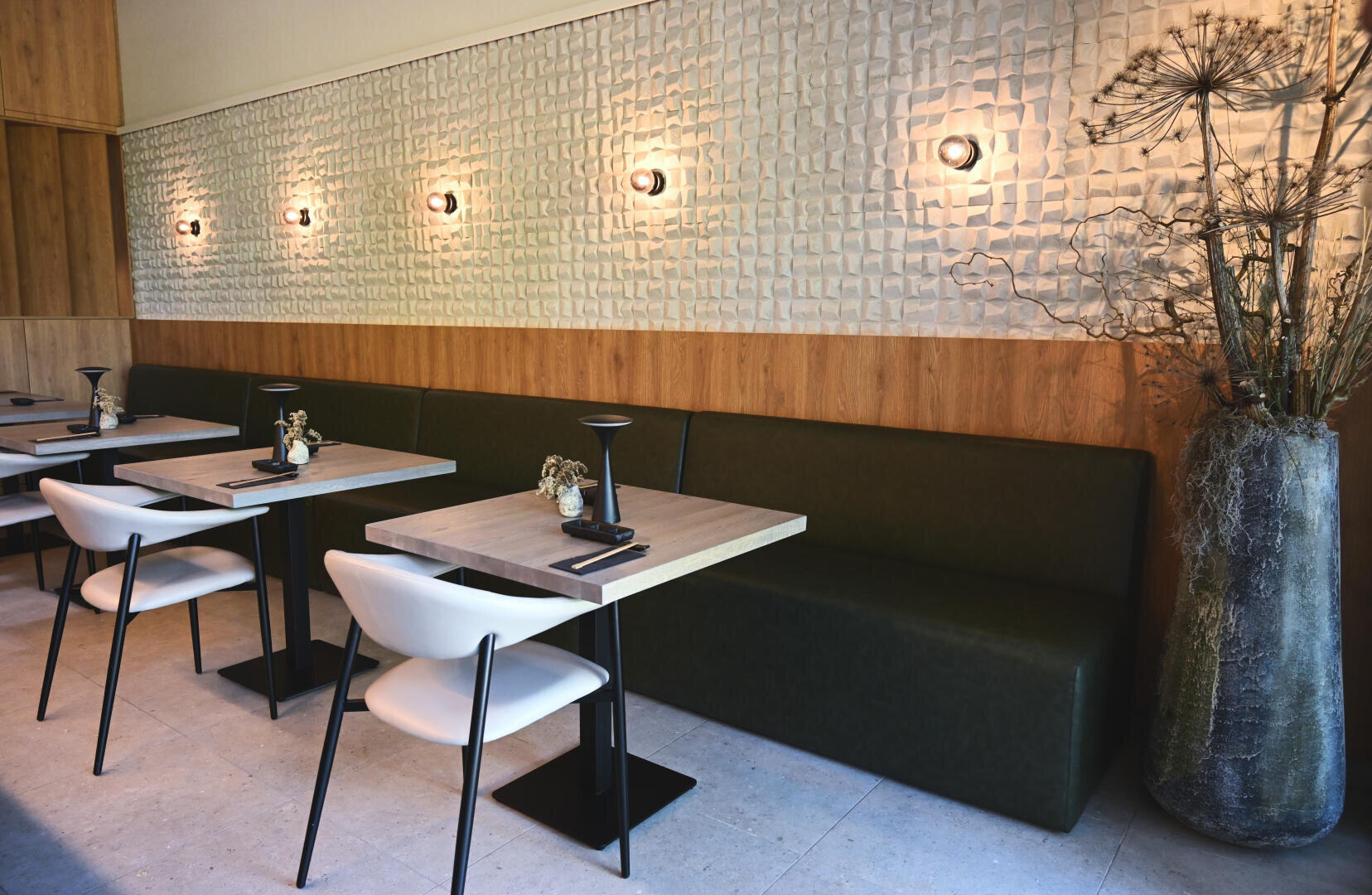 Un intérieur de café moderne avec des tables en bois minimalistes, des chaises blanches, une longue banquette verte rembourrée, des panneaux muraux blancs texturés et un éclairage mural doux. Un grand vase avec des plantes séchées se trouve dans un coin.