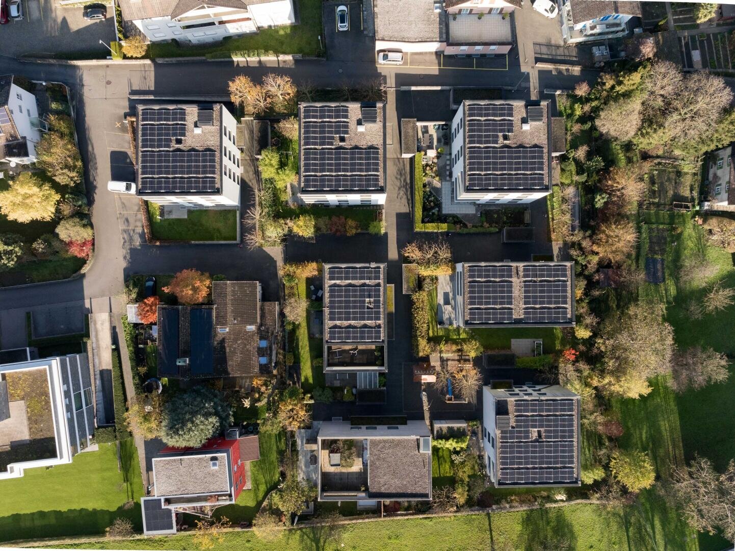 Luftaufnahme von mehreren modernen Wohngebäuden mit Sonnenkollektoren auf den Dächern, umgeben von Bäumen, Rasenflächen und Straßen in einem Vorstadtviertel.