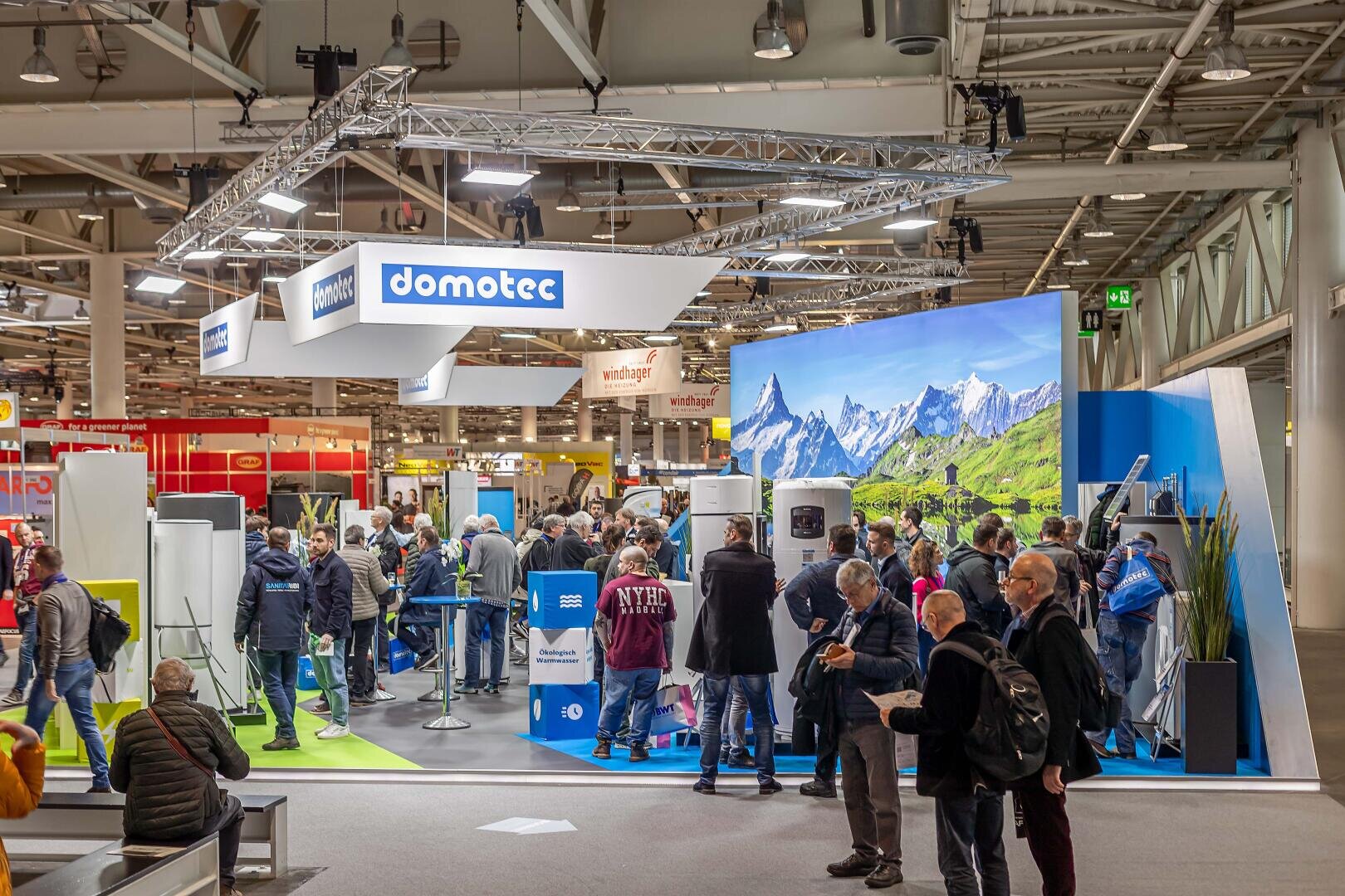Eine Menschenmenge besucht den domotec-Ausstellungsstand auf einer Fachmesse. Der Stand verfügt über große Produktdisplays und eine Kulisse mit einer malerischen Berglandschaft. Der Innenraum ist hell beleuchtet und industriell dekoriert.