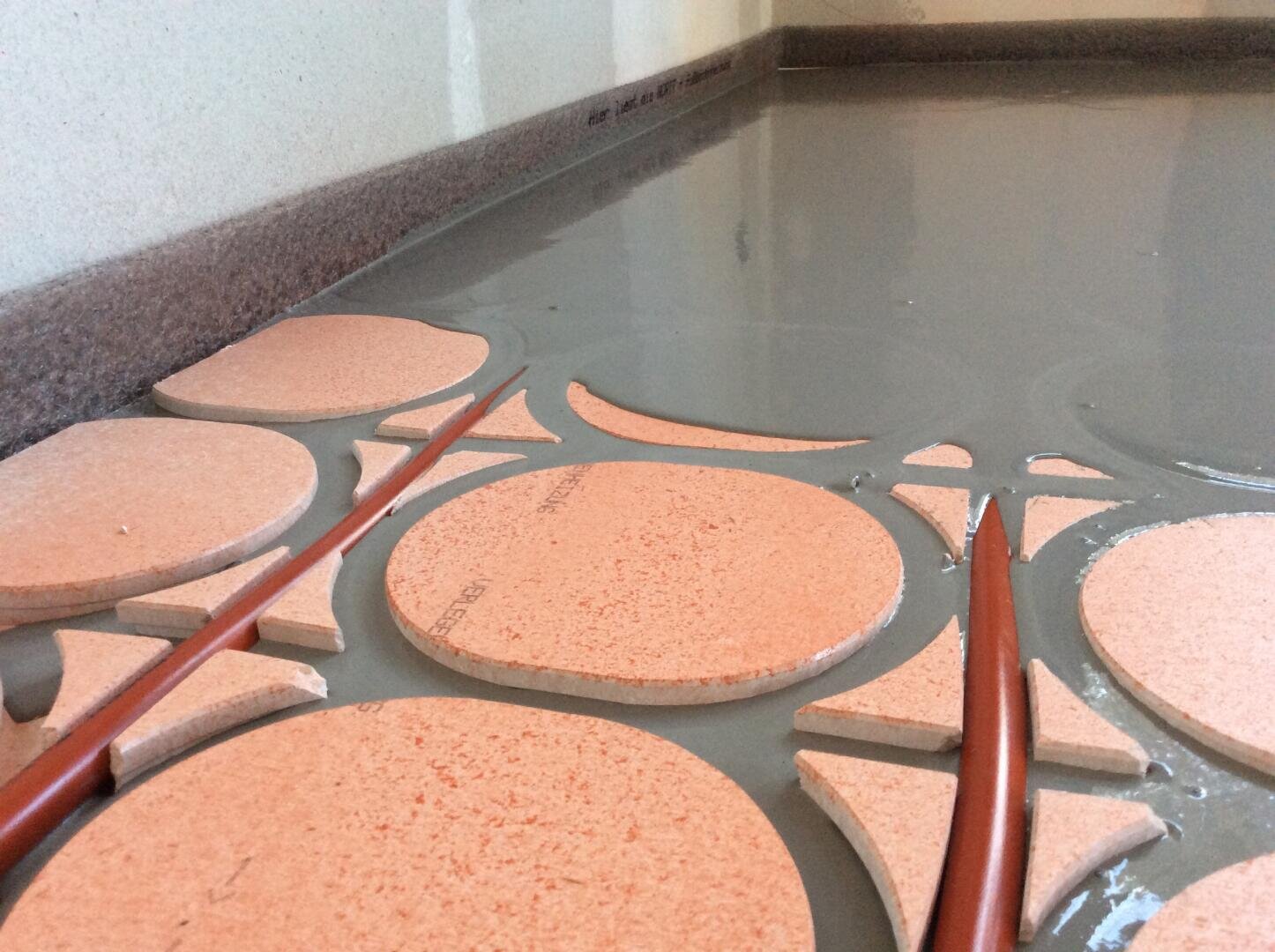 Nahaufnahme eines Fußbodens mit runden und gebogenen orangefarbenen Dämmplatten und roten Schläuchen, die mit einer Schicht frisch gegossenem grauen Fließestrich bedeckt werden. Die Szene zeigt eine Fußbodenheizung, die gerade verlegt wird.