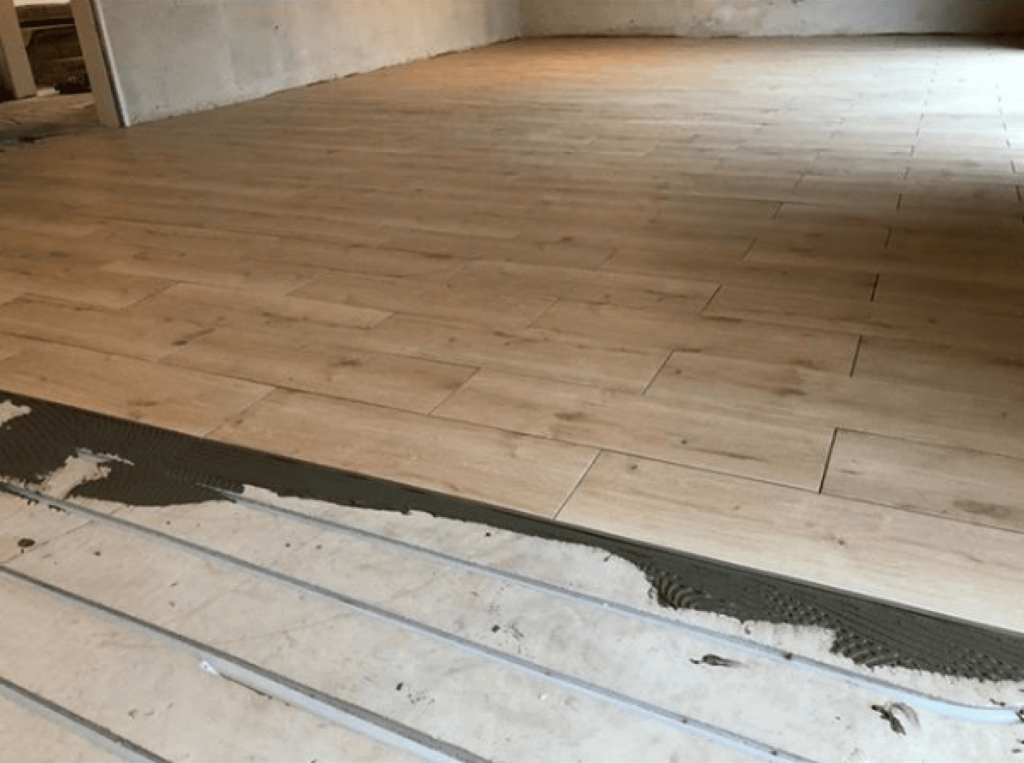 Ein Raum, in dem Keramikfliesen in Holzoptik verlegt werden; der Kleber ist auf dem Boden verteilt, und die Fliesen sind teilweise verlegt, mit sichtbaren Lücken, wo die Verlegung noch nicht abgeschlossen ist.