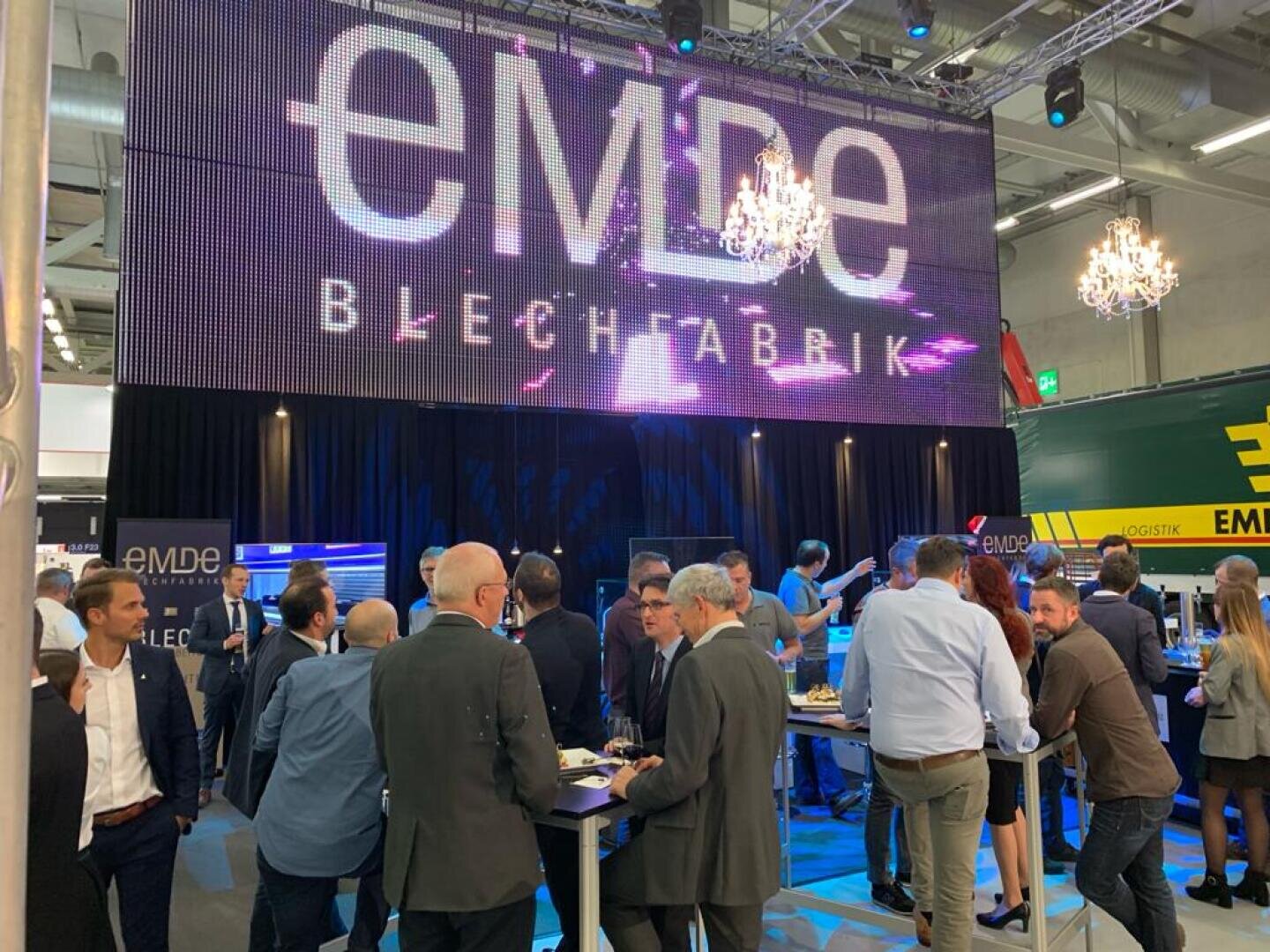 Eine große Gruppe von Menschen in Geschäftskleidung versammelt sich bei einer Indoor-Veranstaltung mit einer großen Leinwand, auf der "EMDE BLECHFABRIK" angezeigt wird. Darüber hängen Kronleuchter, und im Hintergrund sind Informationsstände zu sehen.