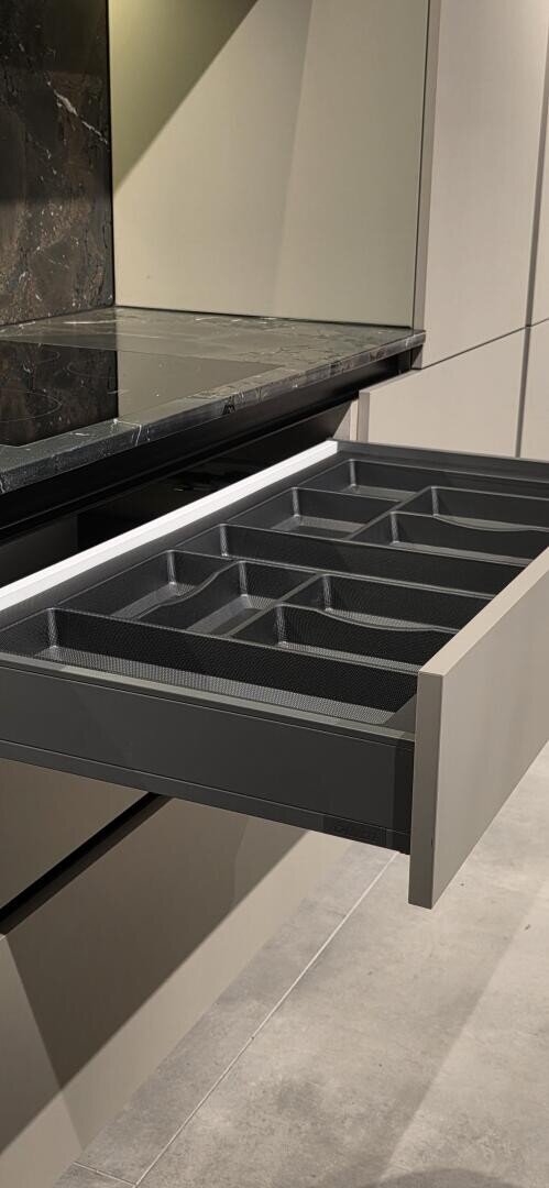 Un tiroir de cuisine ouvert avec plusieurs compartiments noirs vides pour ranger les ustensiles, sous un comptoir en marbre foncé.